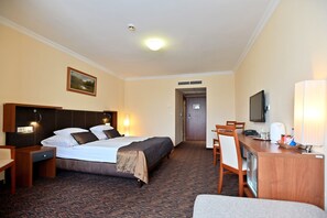 Quarto de hotel com cama, escrivaninha, cadeira, televisão e luminária.