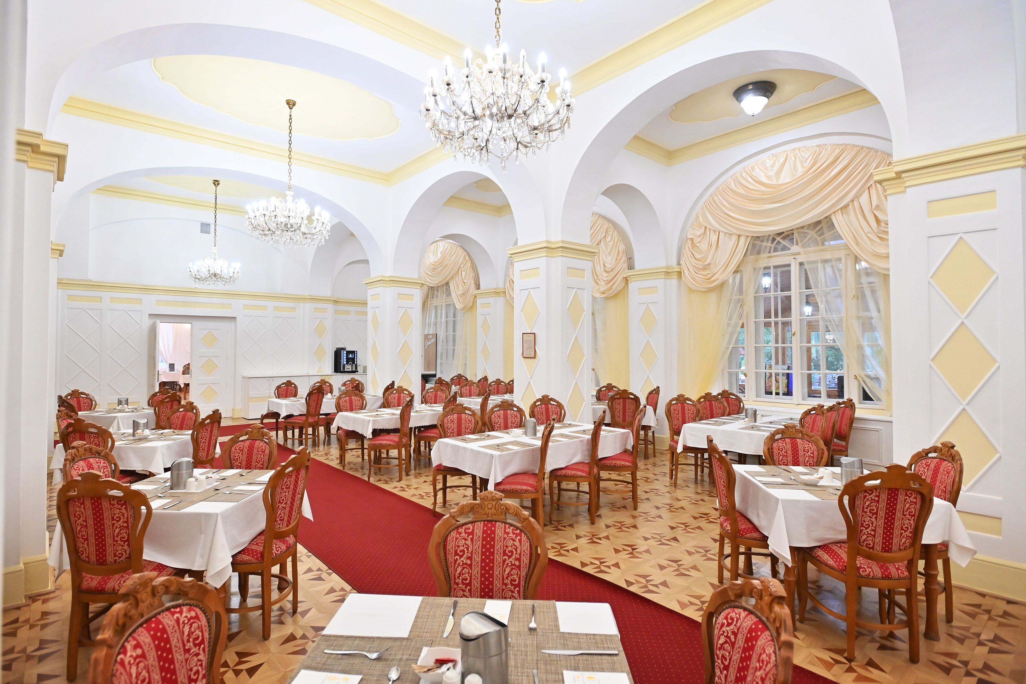 A banquet hall with round tables, red upholstered chairs, chandeliers, and a patterned carpet.