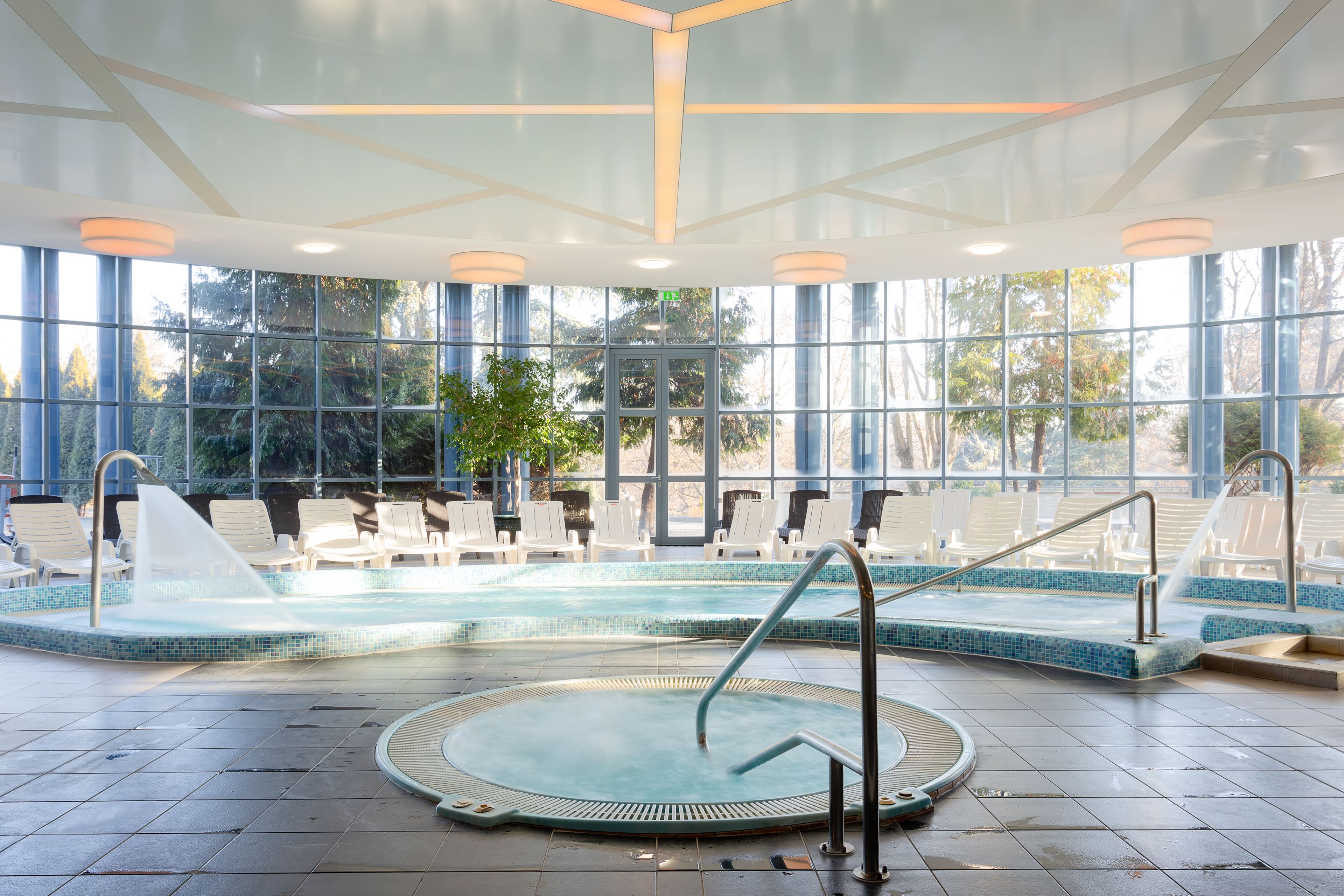 Un espace piscine comprenant un jacuzzi, des chaises longues et une vue sur les arbres à travers de grandes fenêtres.