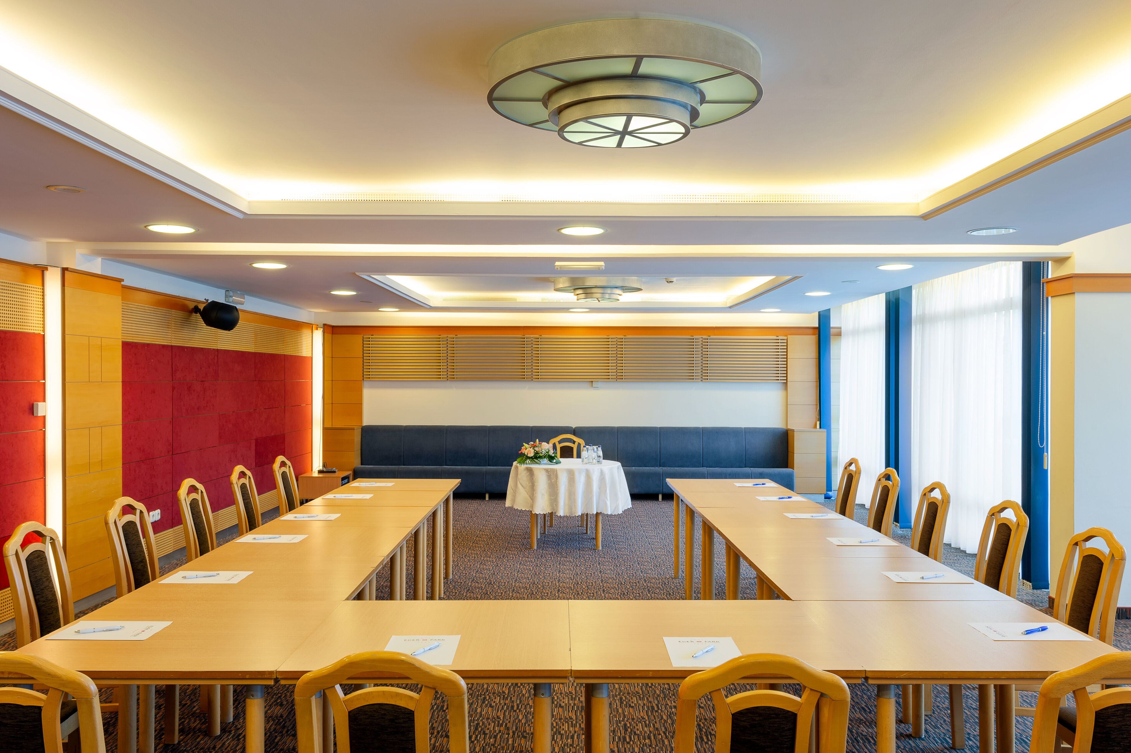 Un salón de conferencias con mesas largas y sillas, una mesa central con arreglo floral, e iluminación empotrada en el techo y una lámpara circular.