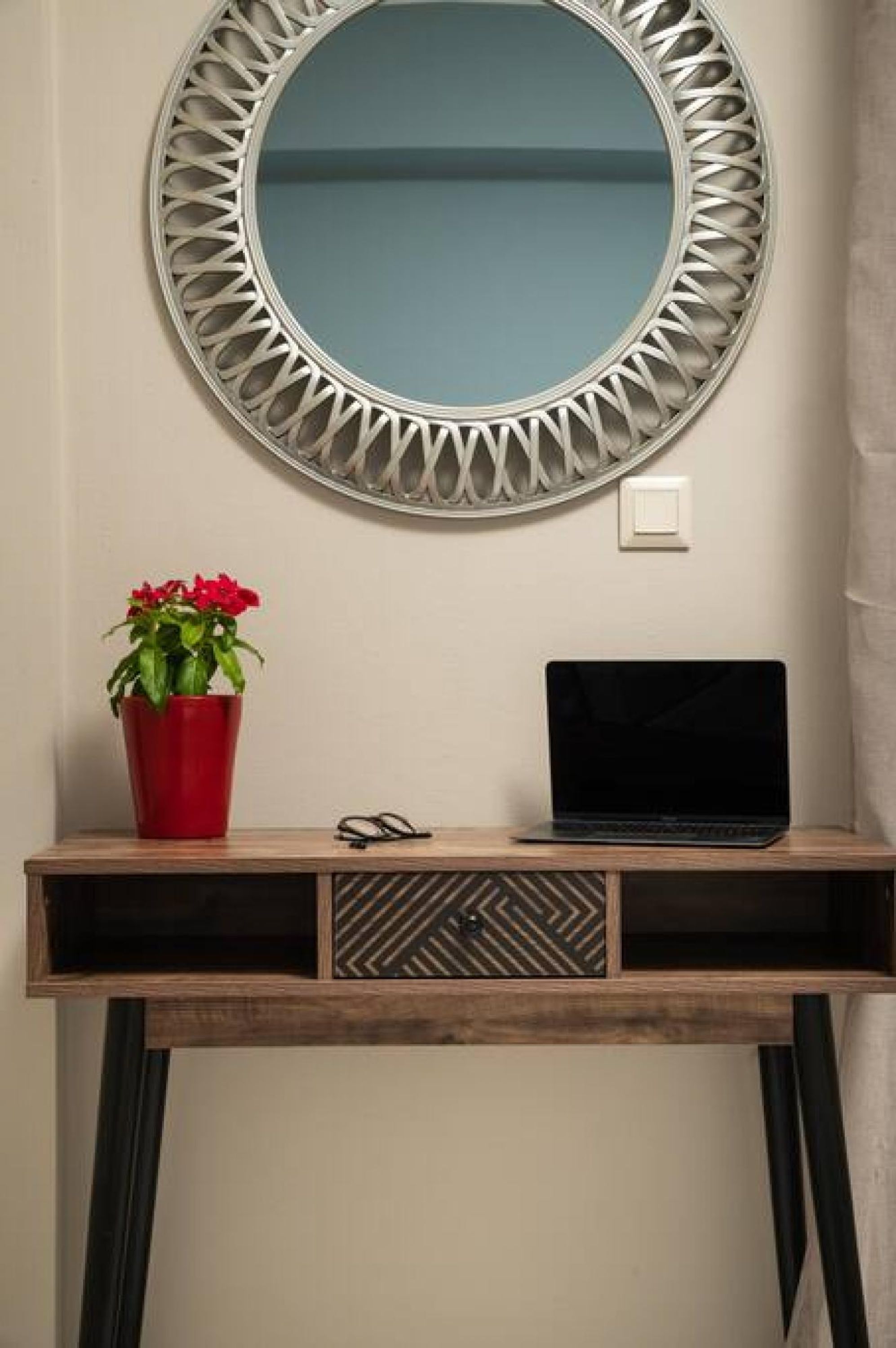 A round mirror with a geometric frame on a wall, a red potted plant, and a laptop on a wooden console table.