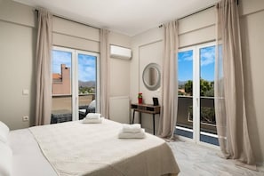A hotel room with a large bed, a round mirror, a small table with a laptop, and a view of the outdoors through large windows.