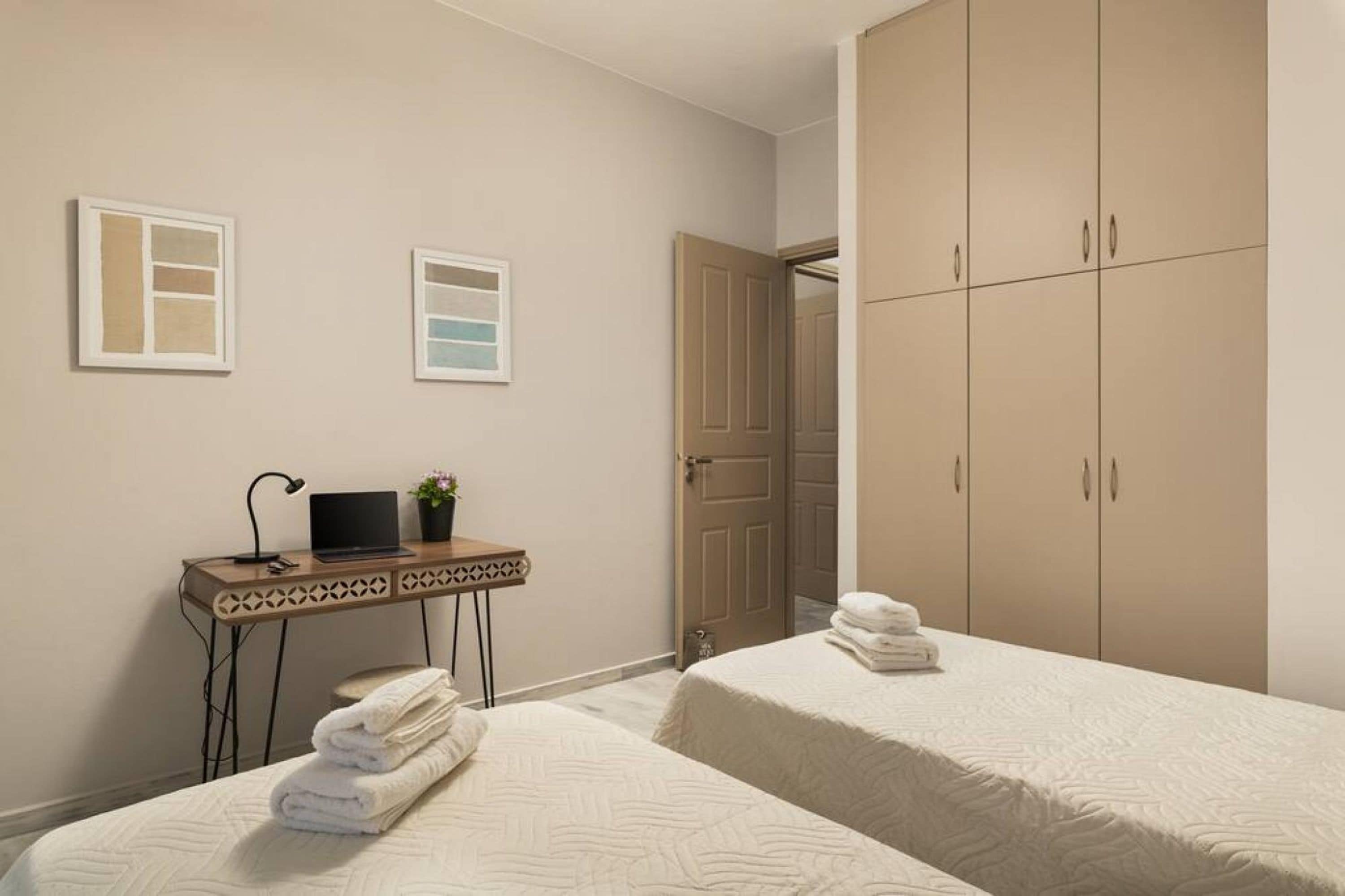 A hotel room with two beds, a desk with a laptop, a small table with a lamp, and a wardrobe.