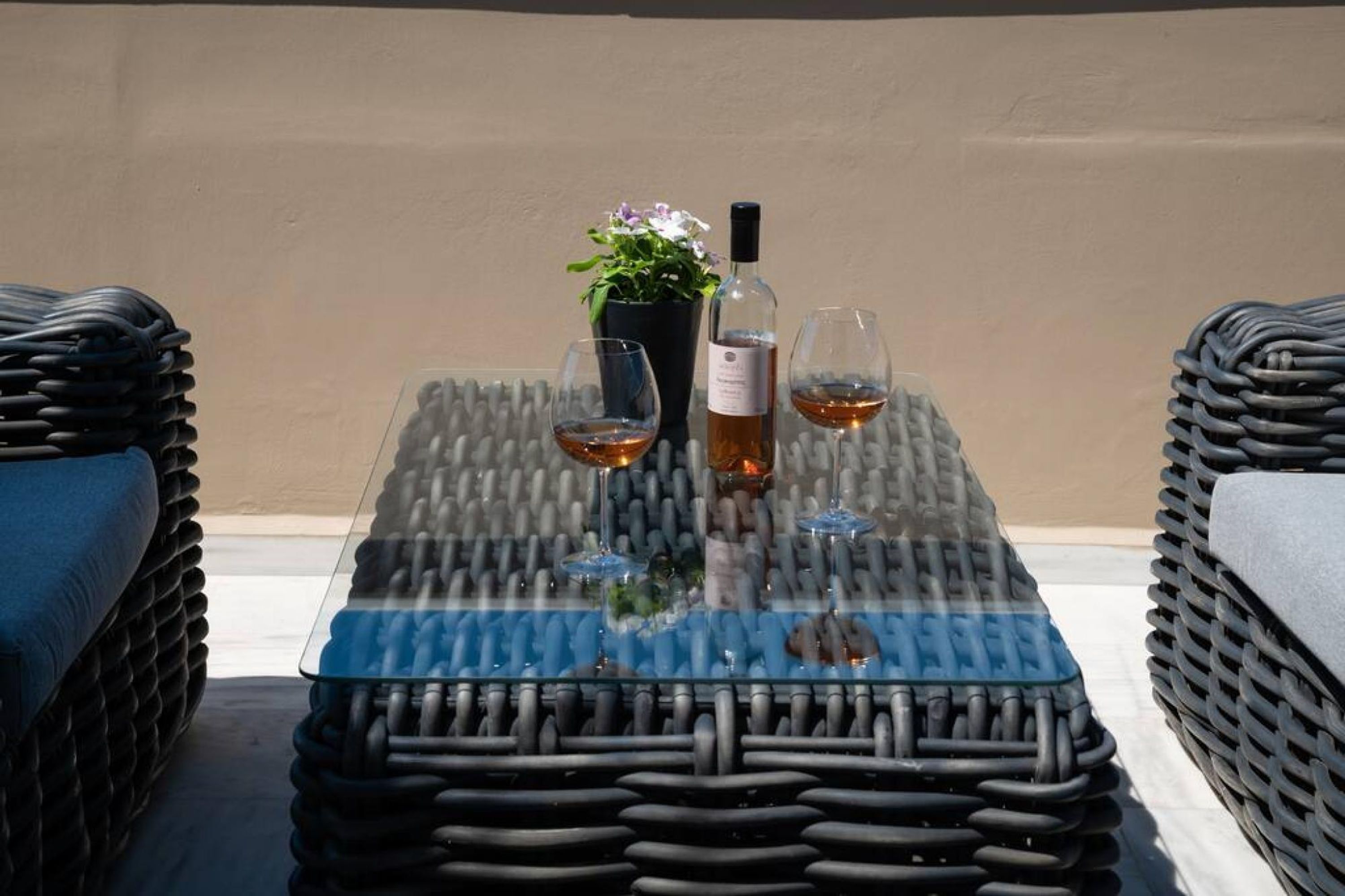 A glass-top table with a potted plant and a bottle, two glasses with a beverage, and wicker chairs.