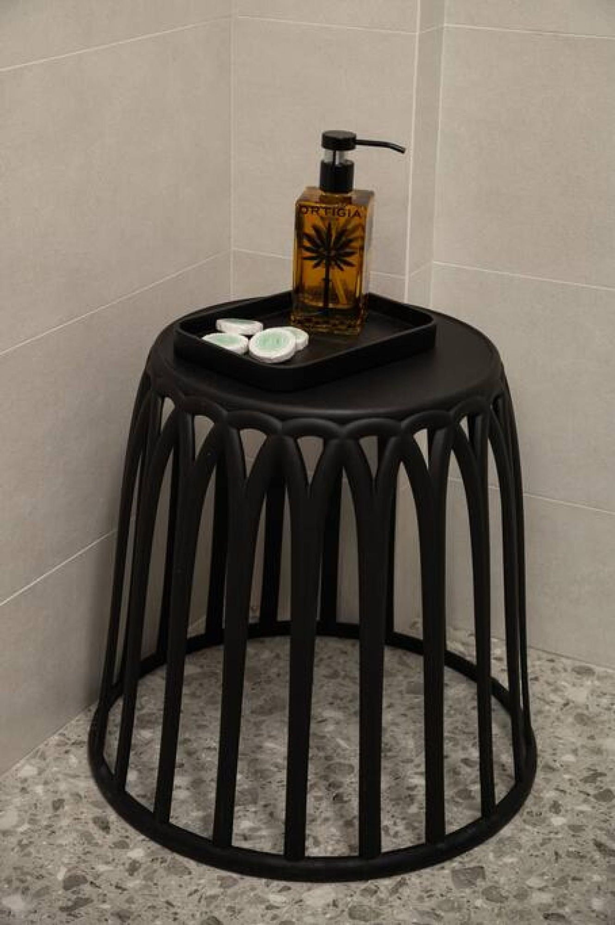 A black stool with a tray and a bottle of lotion against a tiled wall.