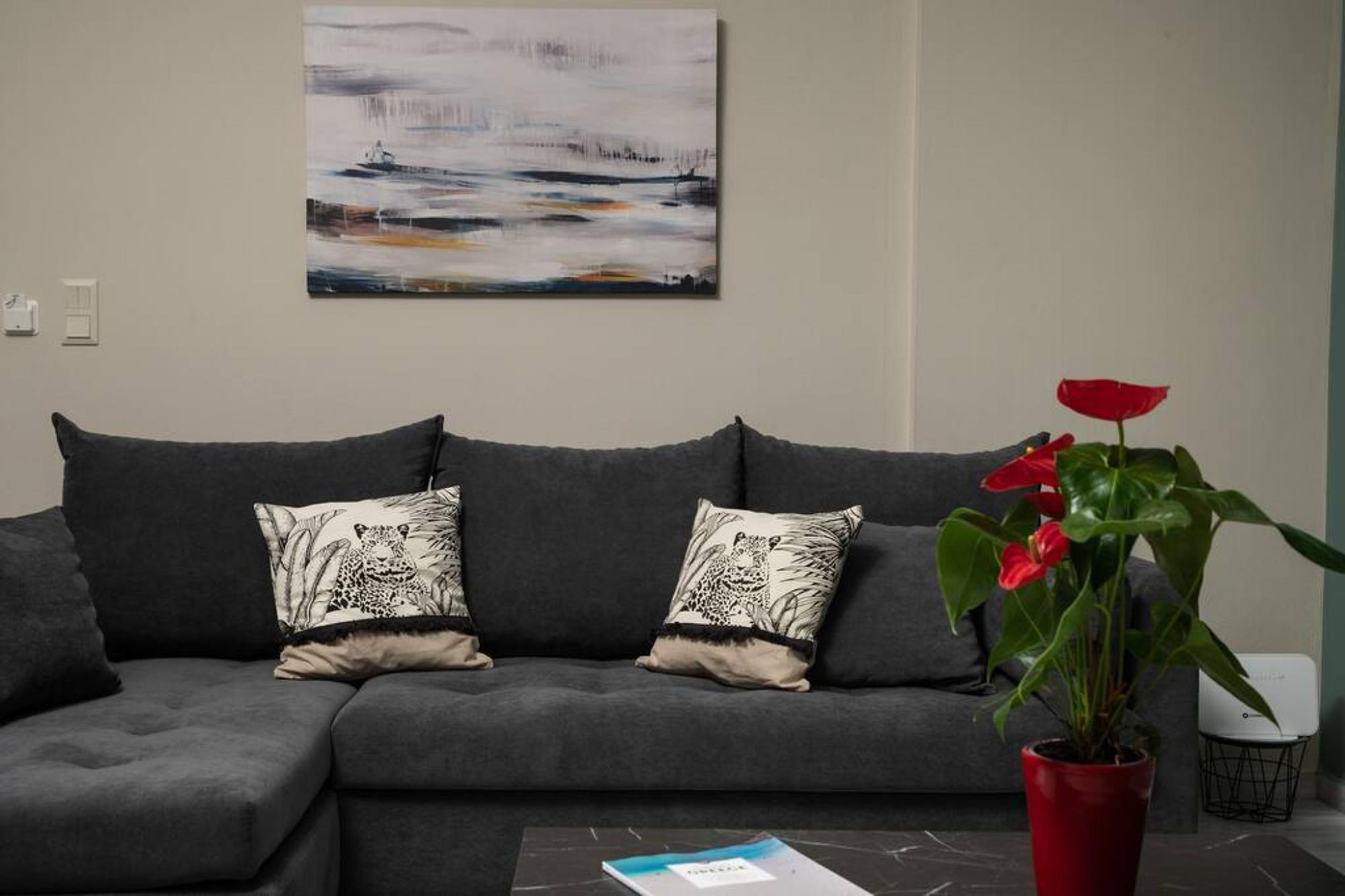 A grey sofa with patterned cushions, a red potted plant, and a painting on the wall.