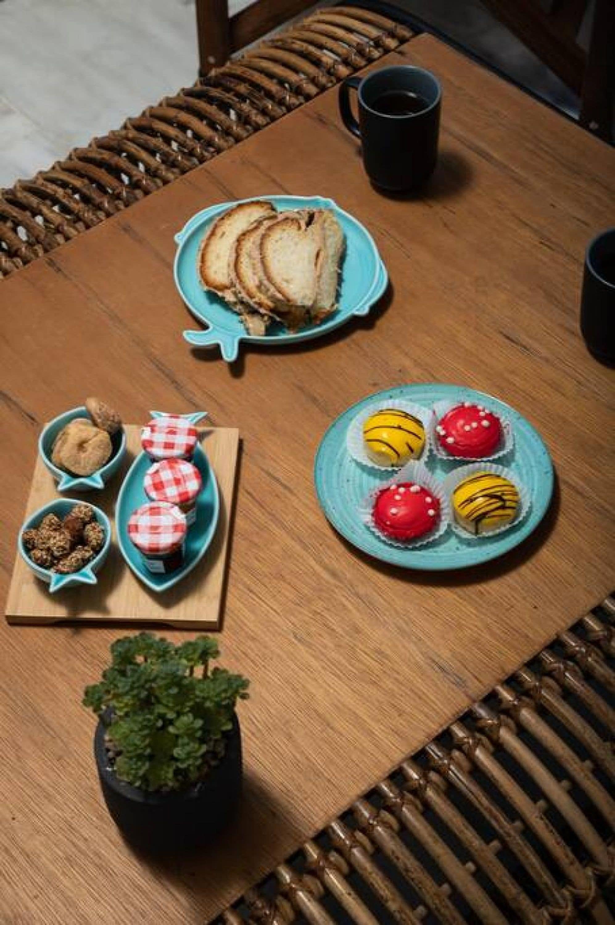 Una mesa de madera con un plato de pan en rodajas, un plato de cupcakes decorados, un cuenco pequeño de frutos secos y una planta en maceta.