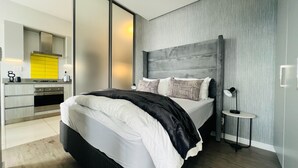 A modern bedroom with a large bed, a bedside table, a lamp, and a view of a kitchen area. - Modern 1 Bed Apartment in Trendy Rosebank (Johannesburg)