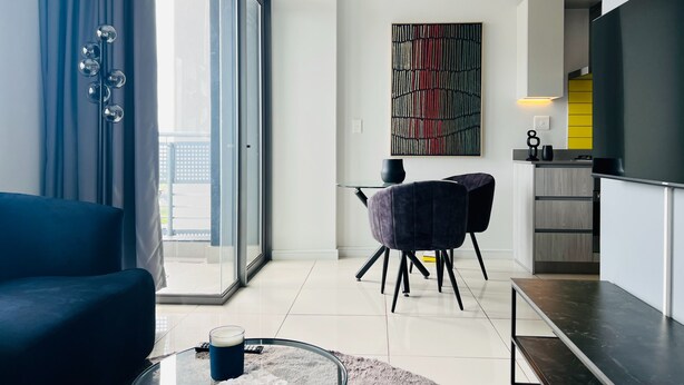 A modern living room with a sofa, a glass coffee table, and a dining area with chairs. - Modern 1 Bed Apartment in Trendy Rosebank (Johannesburg)