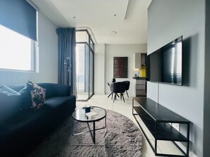 Uma sala de estar moderna com um sofá, uma mesa de centro e uma televisão. Há uma porta de vidro deslizante que dá para uma varanda.