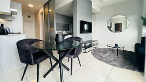A modern living room with a glass dining table, dark armchairs, a TV, and a round mirror. - Modern 1 Bed Apartment in Trendy Rosebank (Johannesburg)