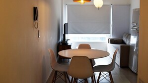 A compact dining area with a round table and four chairs, a hanging pendant light, a wall-mounted telephone, and a window with blinds. - Excelente Ubicación (Ushuaia)