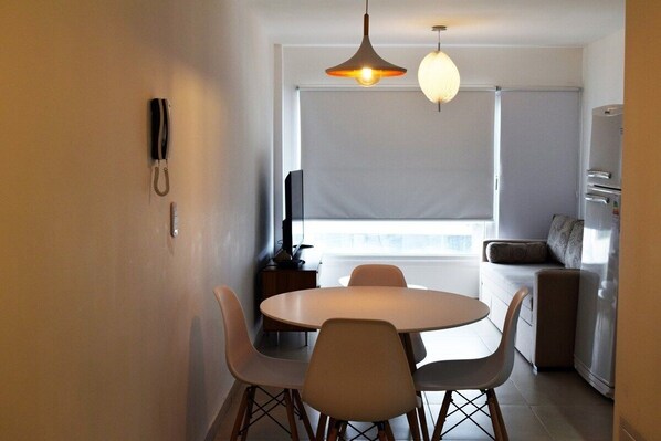 A compact dining area with a round table and four chairs, a hanging pendant light, a wall-mounted telephone, and a window with blinds.