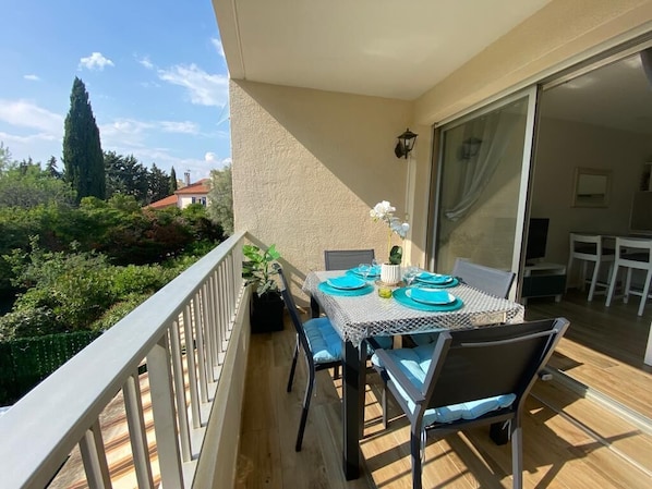 A balcony with a table set for two, chairs, a potted plant, and a view of greenery and buildings.