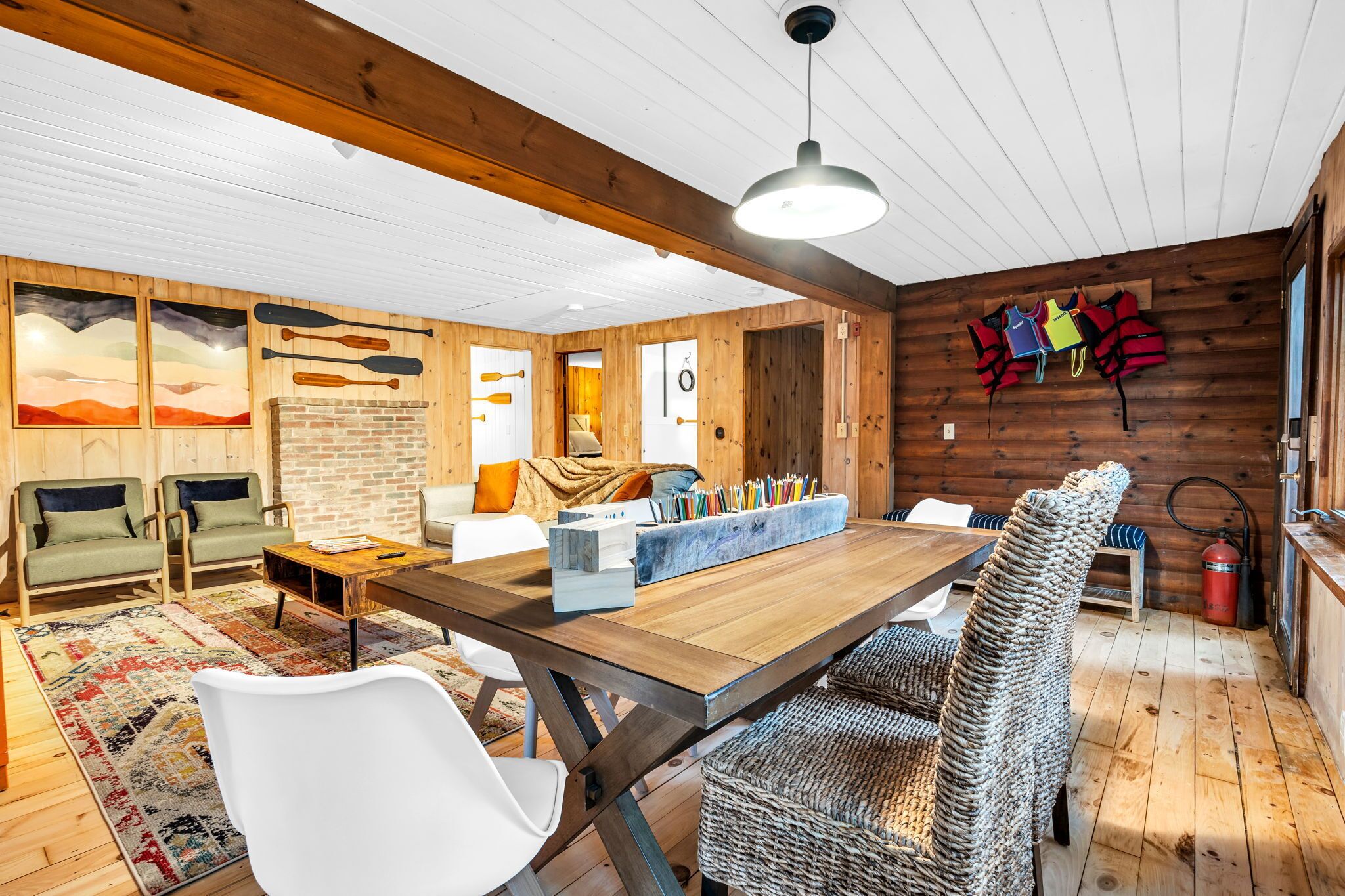 A cozy cabin interior with a wooden dining table, white chairs, a fireplace, and a mounted wall of paddles.