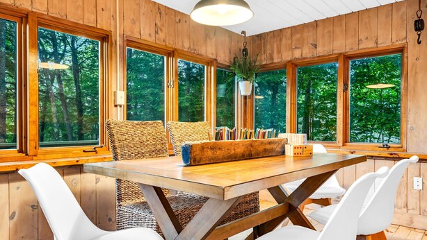 A wooden interior with a large table, white chairs, and large windows offering a view of trees. - Pine and Eagle- Family & Dog Friendly Great Views (Sebago)