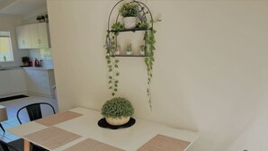 Un coin repas avec une table dressée pour quatre personnes, entourée de chaises. On peut voir une petite plante en pot sur la table et une étagère fixée au mur, où sont disposées d’autres plantes.