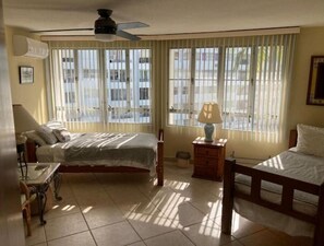 Ein Zimmer mit zwei Betten, einem Deckenventilator, einem Nachttisch mit Lampe und großen Fenstern mit Jalousien.
