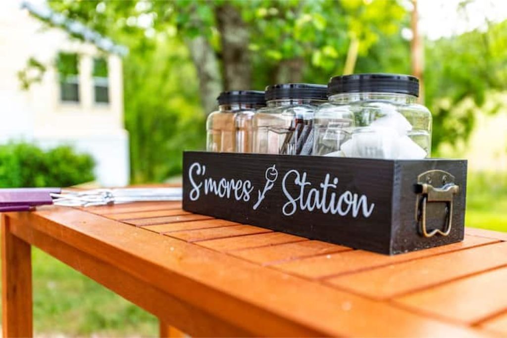A wooden table with a black box labeled "S'mores Station" containing jars of marshmallows and chocolate.