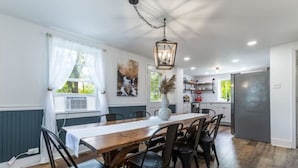 A dining area with a wooden table and black chairs, a hanging light fixture, and a view of the kitchen in the background.