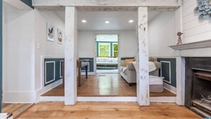 A room with a wooden floor, a fireplace, and a view into another room with a bed and a window.
