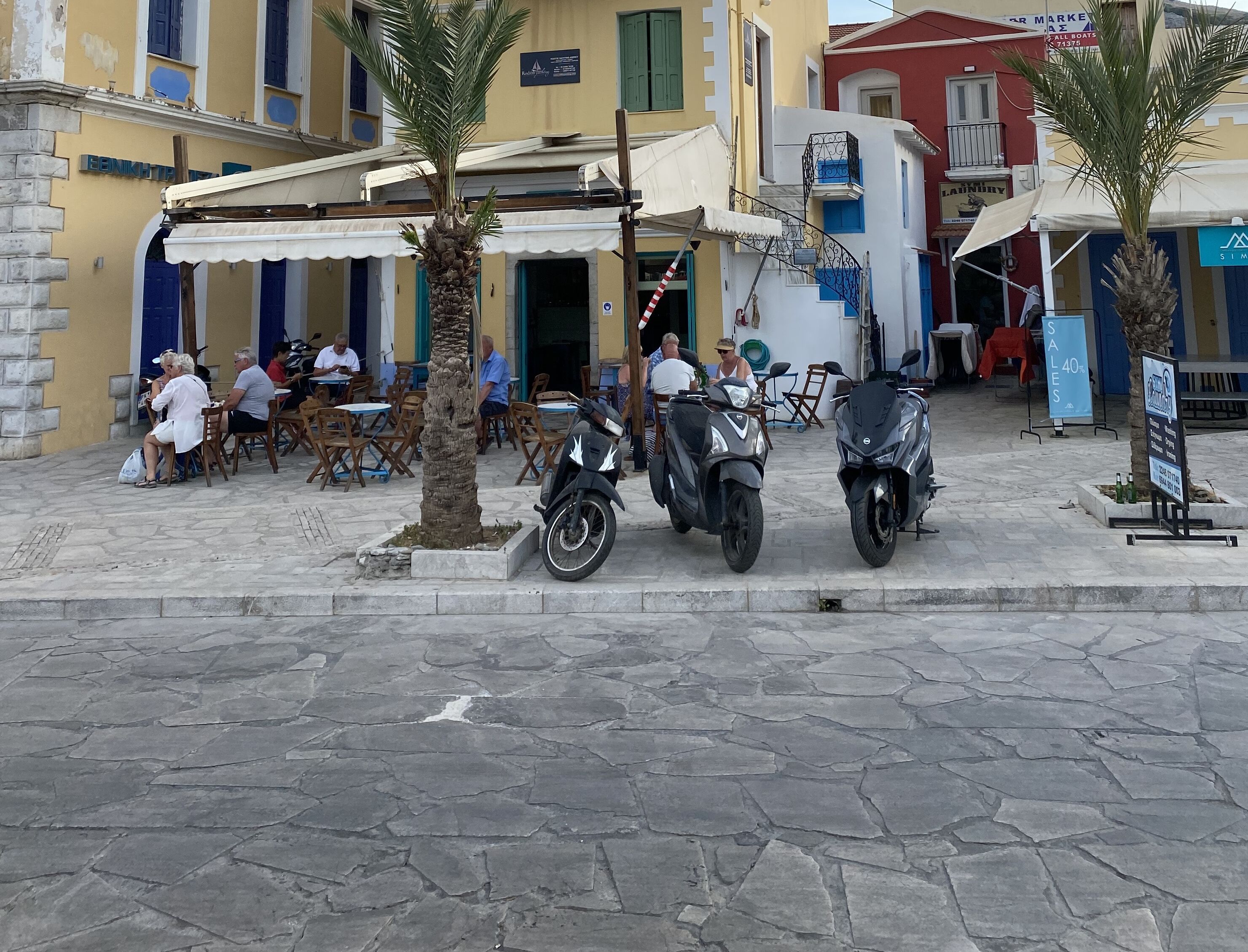 A street with outdoor seating, palm trees, and parked scooters.