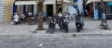 A street with outdoor seating, palm trees, and parked scooters.