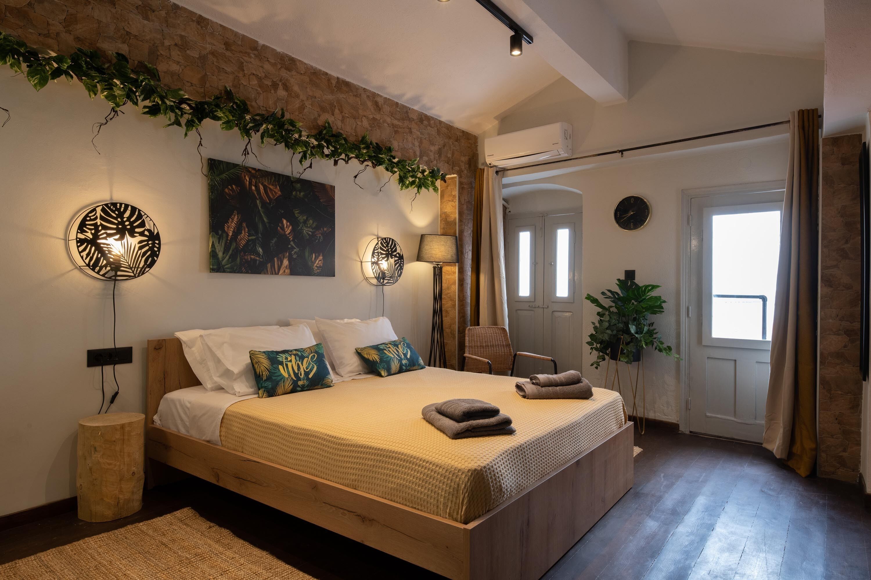 A bedroom with a wooden bed, a wall-mounted lamp, a painting, and a potted plant.