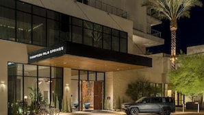 A modern hotel entrance with a sign reading 'Thompson Palm Springs,' a car parked outside, and a palm tree.