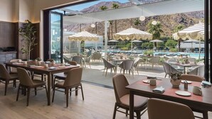 A modern dining area with tables and chairs, a pool with umbrellas, and a mountain view.