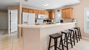 A kitchen with wooden cabinets, a white refrigerator, a microwave, and a coffee maker. There are three stools at the island. - 2 King Beds - Walk to Beach (St. Augustine)