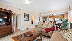 A spacious living room with a large sofa, a wooden coffee table, and a television. A kitchen is visible in the background with wooden cabinets and a chandelier. - 2 King Beds - Walk to Beach (St. Augustine)