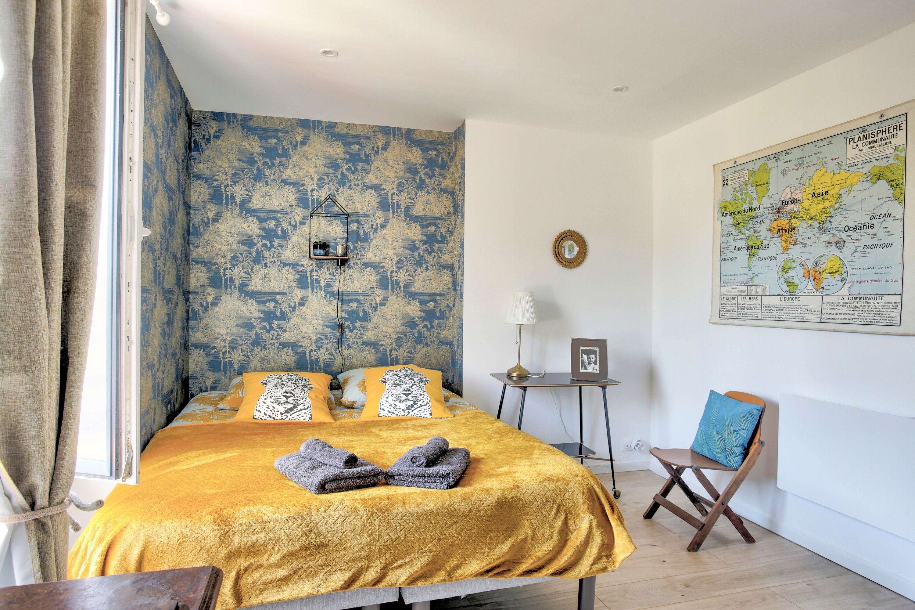 A bedroom with a bed, a map on the wall, a small table with a lamp, and a chair.