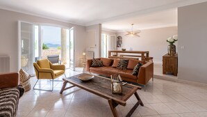 A spacious living area with a sofa, armchairs, and a coffee table. There is a dining table with chairs, a bookshelf, and a view of the outdoors.