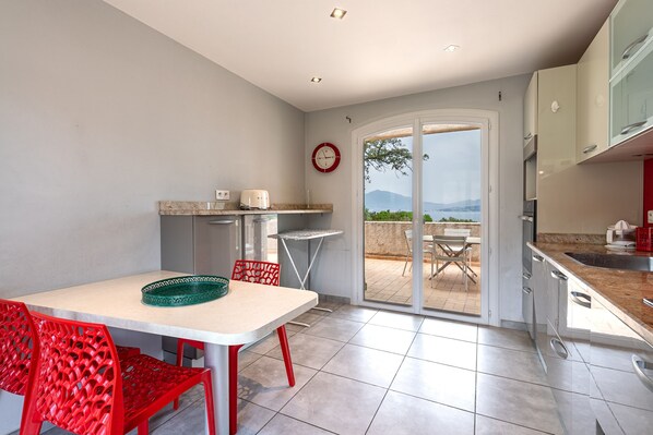 A modern kitchen with a dining area, featuring a table with red chairs, a clock on the wall, and a view through the glass door.