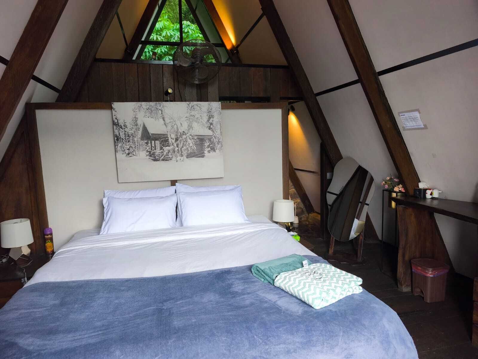 A bedroom with a bed, two bedside lamps, a fan, a desk with a chair, and a painting on the wall.