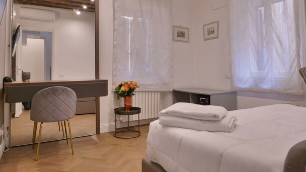 Een slaapkamer met een bed, een stoel, een klein tafeltje met een bloevas en een grote spiegel.