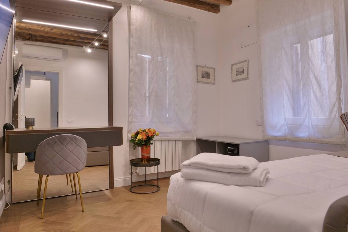 Ein Schlafzimmer mit einem Bett, einem Sessel, einem kleinen Tisch mit Blumenvase und einem großen Spiegel.