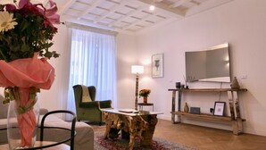 Een woonkamer met een houten salontafel, een groene fauteuil, een bank, een aan de muur bevestigde televisie, en een vaas met bloemen op een bijzettafeltje.