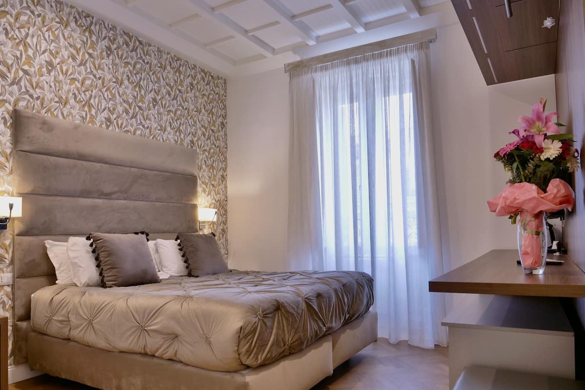Ein Schlafzimmer mit einem großen Bett, einem Kopfteil, einem Nachttisch, einer Lampe, einem Fenster mit durchsichtigen Vorhängen und einer Vase mit Blumen auf einem Tisch.