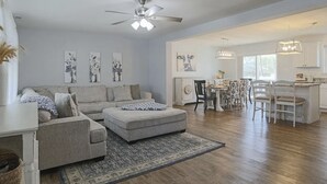 A spacious living room with a large grey sofa, a matching ottoman, and a patterned rug. There is a dining area with a table and chairs, and a kitchen visible in the background.