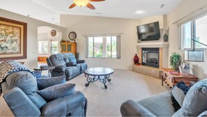A spacious living room with a fireplace, comfortable seating, a ceiling fan, and a large window providing natural light. - Hole in One! Luxury Home Near Golf Course, Airport, Embry Riddle (Prescott)