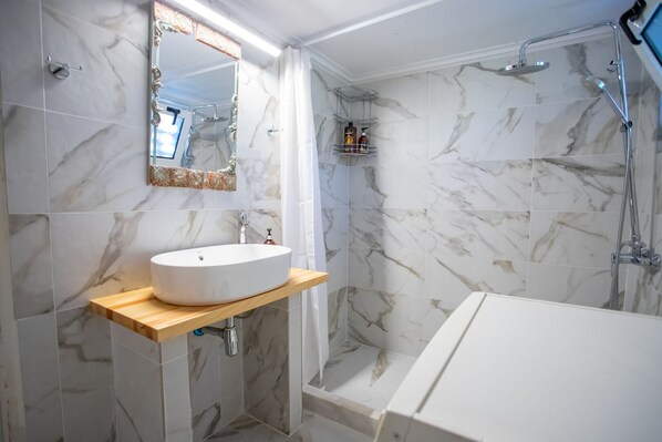 Un baño con ducha revestida de mármol, un lavabo blanco sobre una estantería de madera y un espejo con marco decorativo.