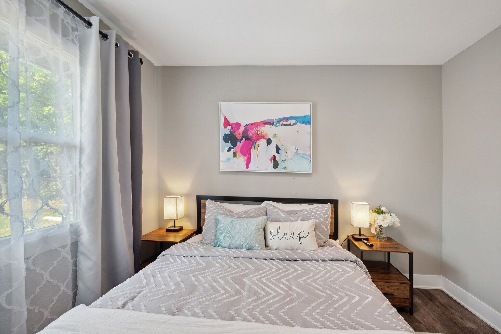 A neatly made bed with a patterned bedspread, two bedside lamps, a nightstand with a vase of flowers, and a colorful abstract painting on the wall.