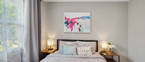 A neatly made bed with a patterned bedspread, two bedside lamps, a nightstand with a vase of flowers, and a colorful abstract painting on the wall.