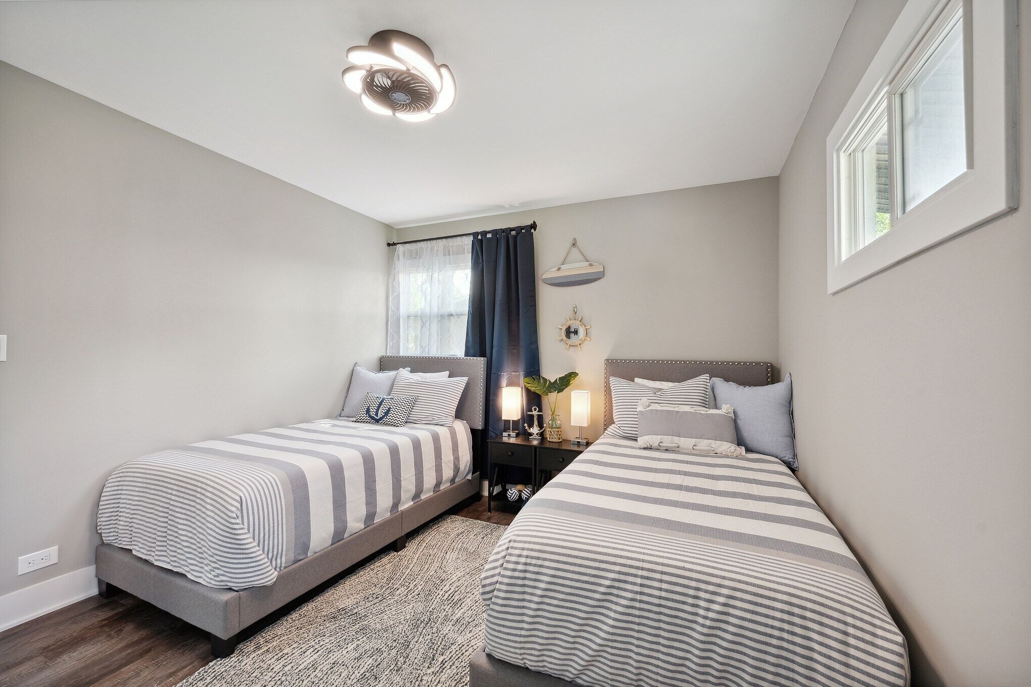 A bedroom with two single beds, a bedside table with lamps, a hanging plant, and a window with curtains.