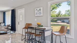 A modern kitchen with a dining area, featuring a large window with a view of the outdoors, a kitchen island with stools, and a door with a decorative handle. - Charming Contemporary Family Home 3 Bedroom Home by RedAwning (Glenview)