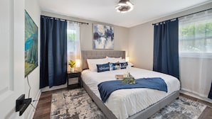 A bedroom with a bed, a nightstand, a lamp, a painting, and a window with curtains. - Charming Contemporary Family Home 3 Bedroom Home by RedAwning (Glenview)