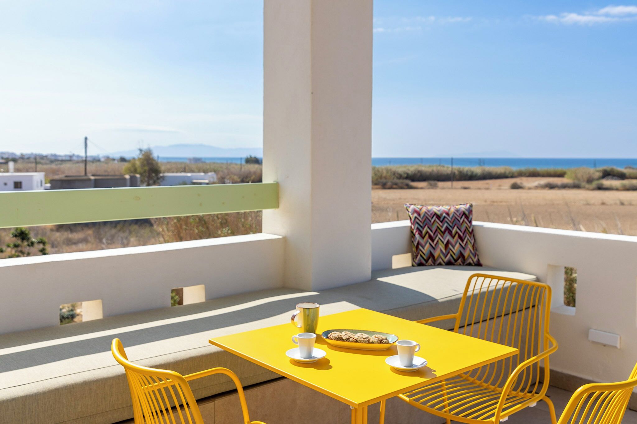Una mesa amarilla con dos tazas y un plato de galletas en un balcón con vista al mar y a la costa.