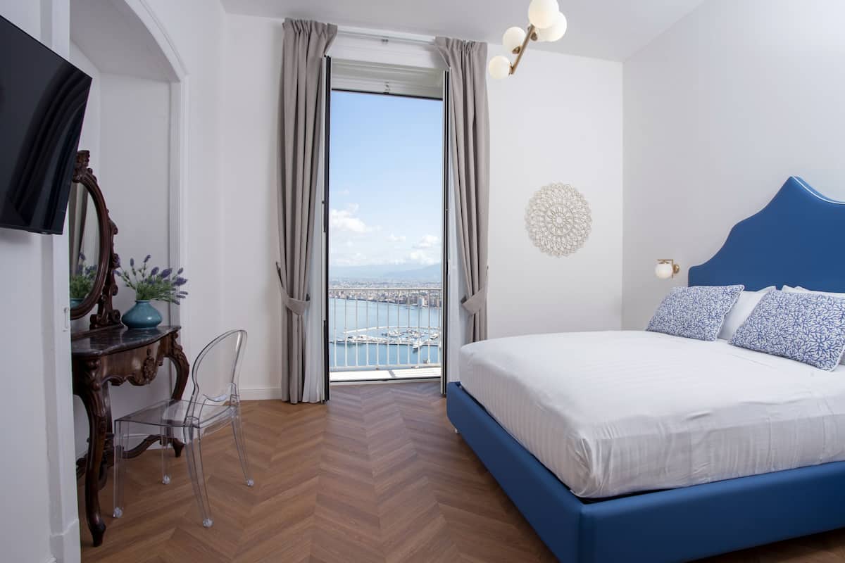 A bedroom with a bed, a chair, a desk, a TV, and a view of the city through a window.